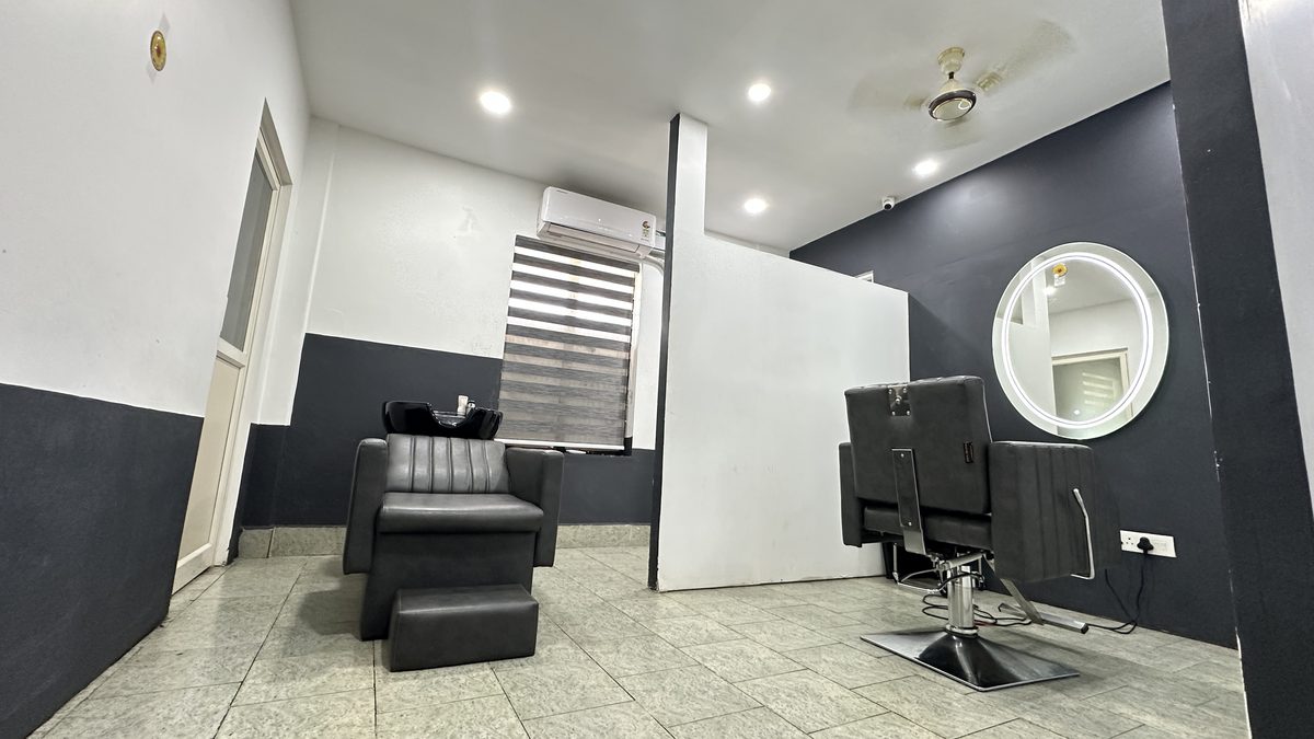 The wash area: a reclining basin chair by a shaded window with an air conditioner above it, and a styling chair with a lit round mirror to the right.