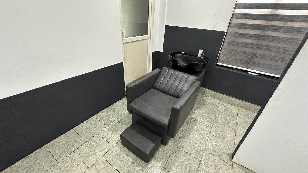 A padded wash chair with a footrest under a black basin, in a corner beside a shaded window.